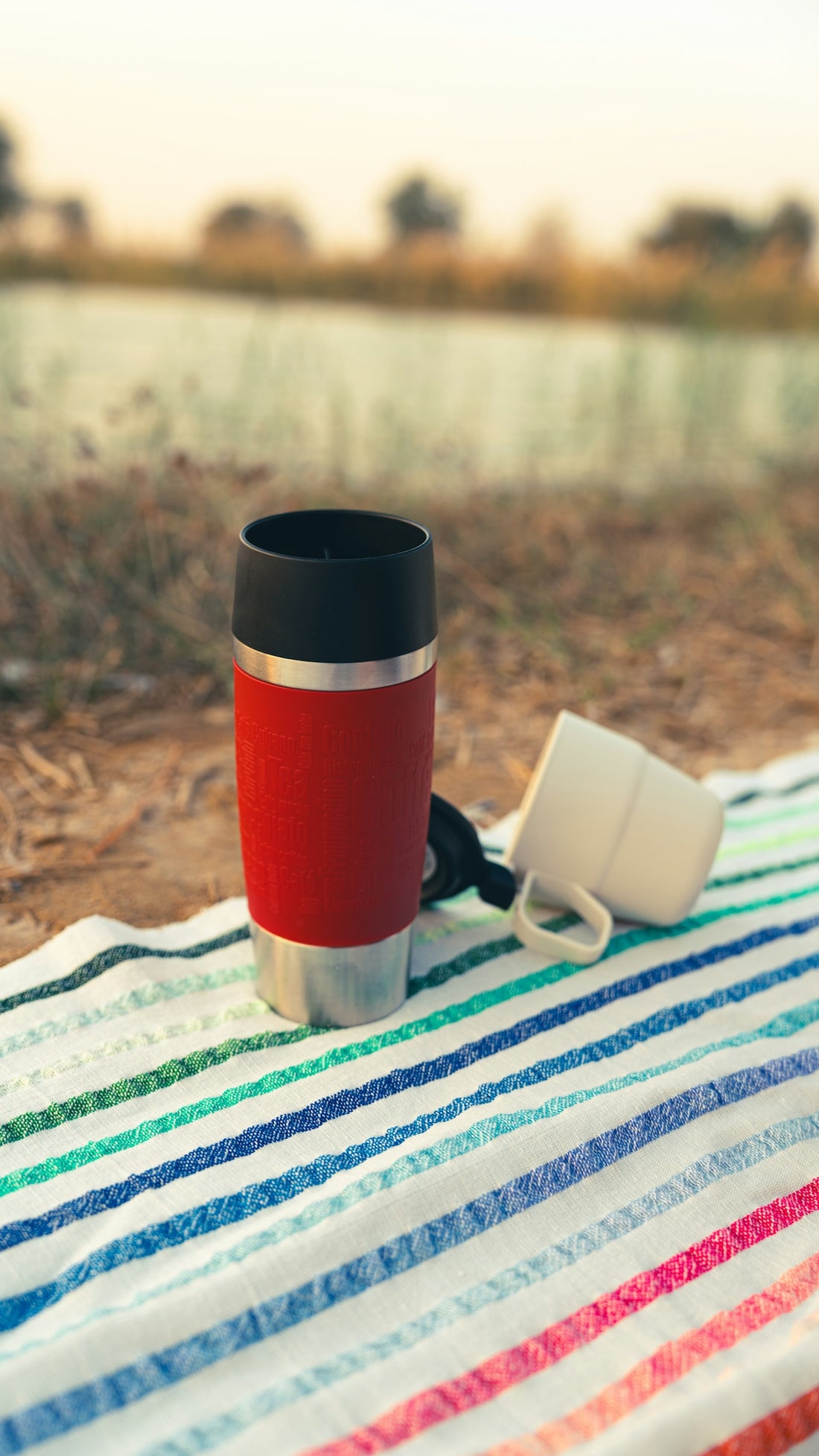 Red thermos and white mug on striped blanket outdoors
